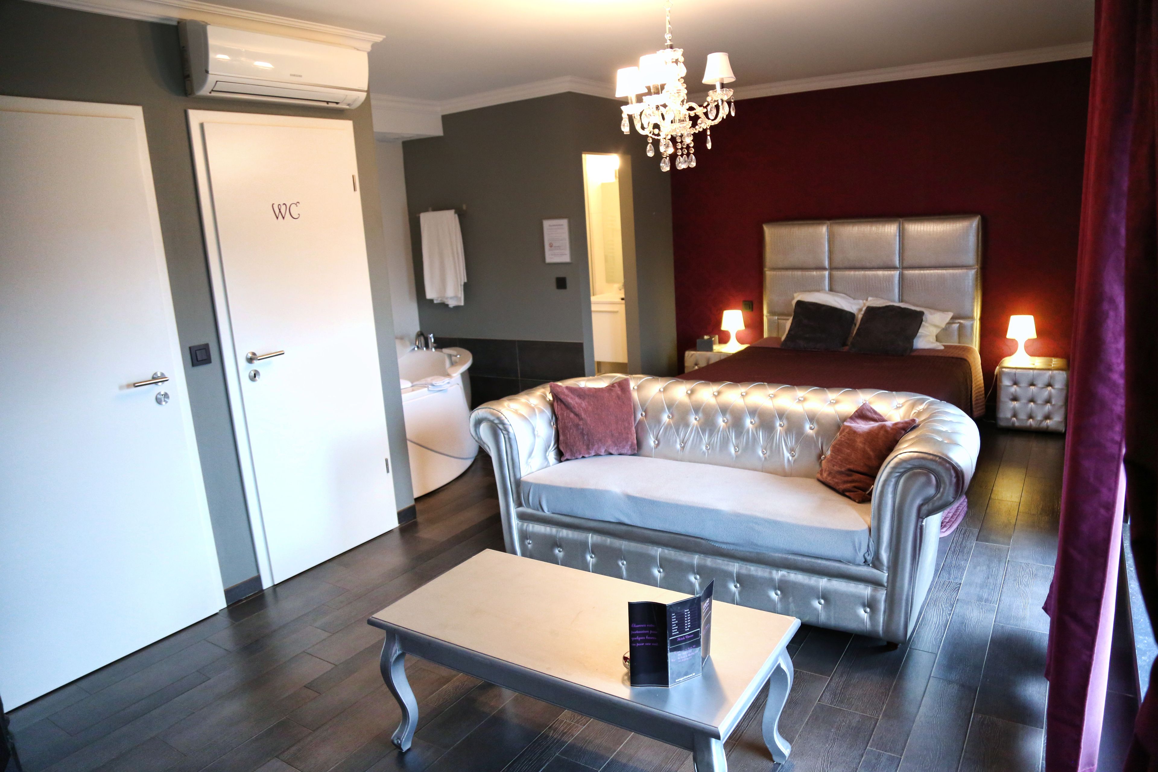 Quarto de hotel com cama, sofá, mesa pequena, lustre, banheiro com pia e porta branca com a inscrição 