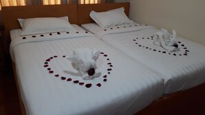 A double bed with white linens, a wooden headboard, and a heart-shaped arrangement of rose petals and a towel on the bed.