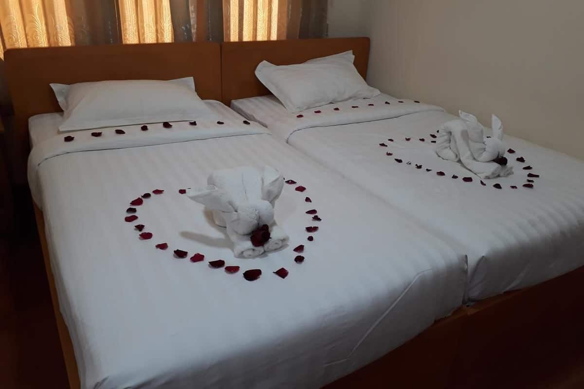 Un lit double avec du linge de lit blanc, une tête de lit en bois et une décoration en forme de cœur composée de pétales de roses, ainsi qu’une serviette posée sur le lit.