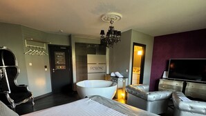 A modern hotel room with a large bed, a sitting area with a sofa, a bathtub, and a television.