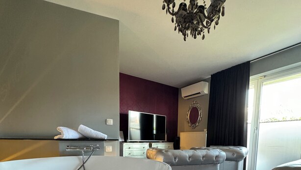 A modern hotel room with a large bathtub, a sofa, a flat-screen TV, and a chandelier.