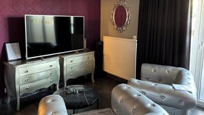 A modern living room with a flat-screen TV, a white sofa, a small round ottoman, a white dresser, and a large mirror on the wall.