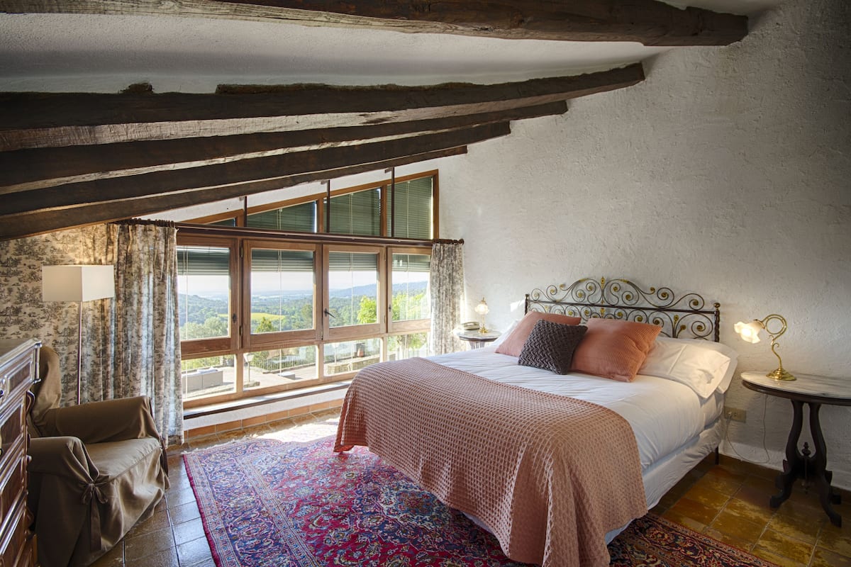A bedroom with a large window, a bed with a patterned bedspread, a nightstand with a lamp, and a wooden chair.