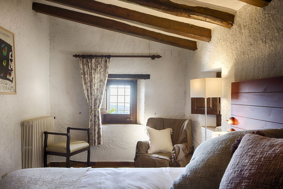 A rustic room with a wooden ceiling, a radiator, a window with curtains, a chair, a bedside table with a lamp, and a bed.