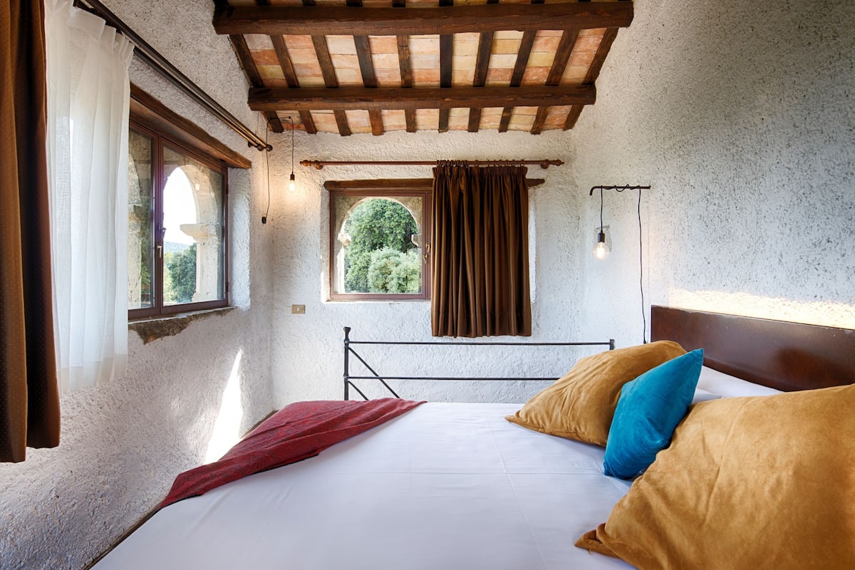A bedroom with a bed, a window with curtains, a wooden ceiling, and a view of greenery outside.