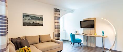 A modern living room with a sofa, a coffee table with a vase of flowers, a blue chair, a desk with a flat-screen TV, and a painting on the wall.