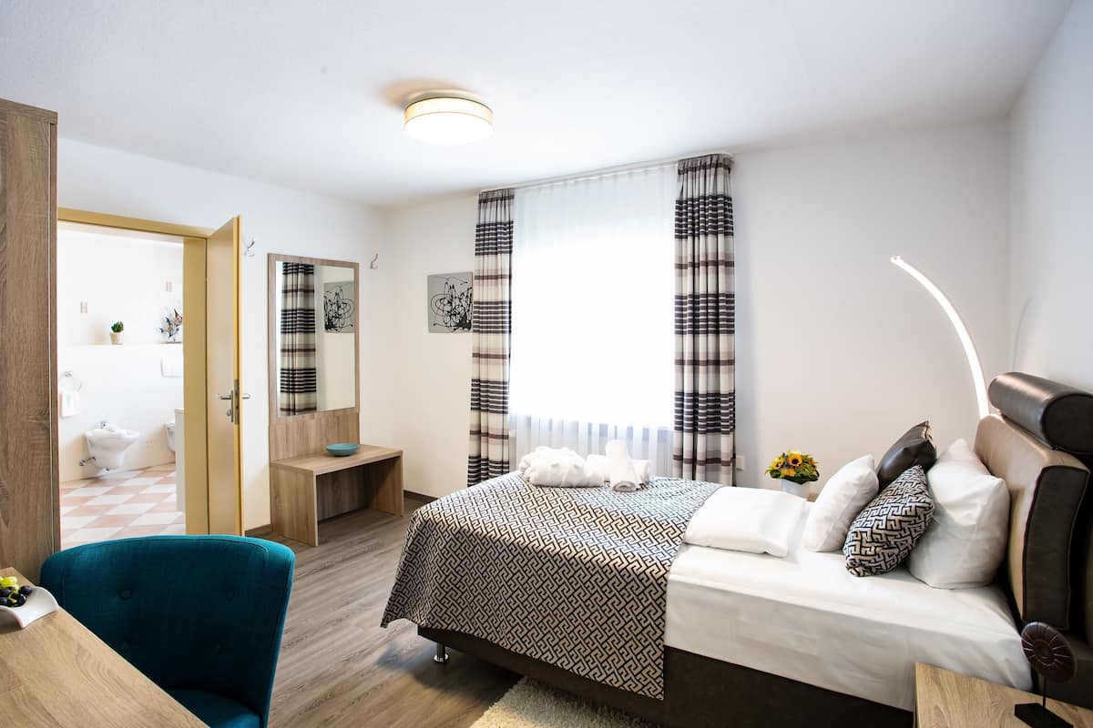 Une chambre d’hôtel moderne comprenant un lit, un bureau avec une chaise, une petite table avec des fruits, et une salle de bain visible à travers une porte ouverte.