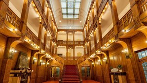 Een statige hal met een centrale trap, versierde houten balustrades en een groot glas-in-loodraam aan de bovenkant.