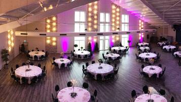 A spacious hall with multiple round tables, each set with chairs and floral centerpieces, under a high ceiling with exposed beams and large windows.