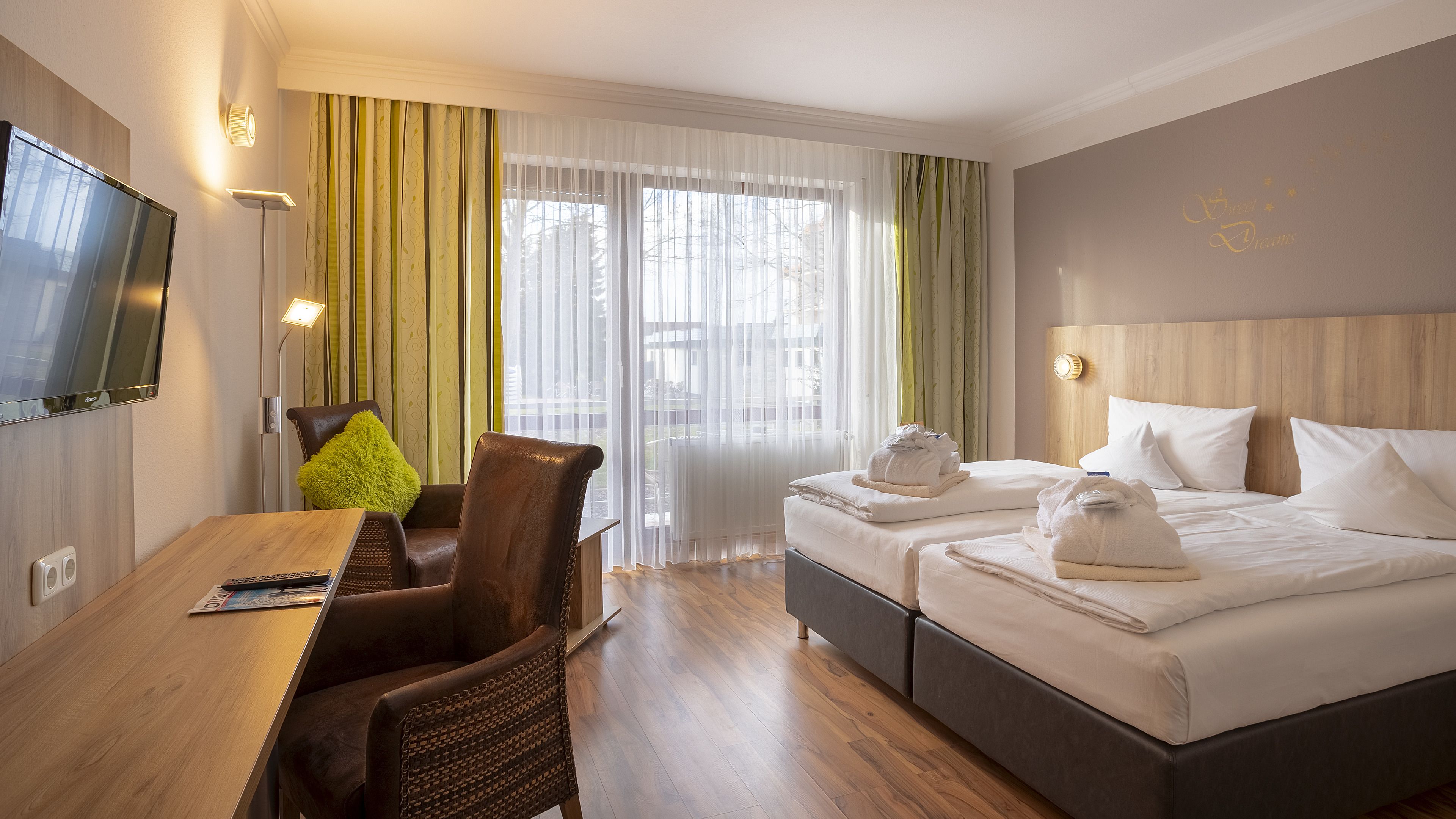 Une chambre d’hôtel avec un bureau en bois, deux lits avec des serviettes, une télévision et une fenêtre avec des rideaux légers.