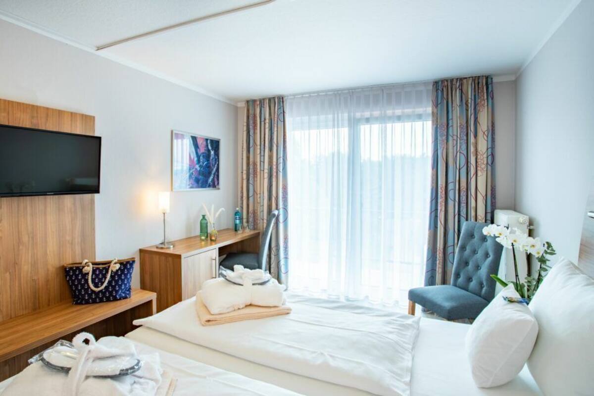 Ein Hotelzimmer mit einem Bett, einer Sitzecke mit einem Sessel, einem an der Wand montierten Fernseher und einem Fenster mit Vorhängen.