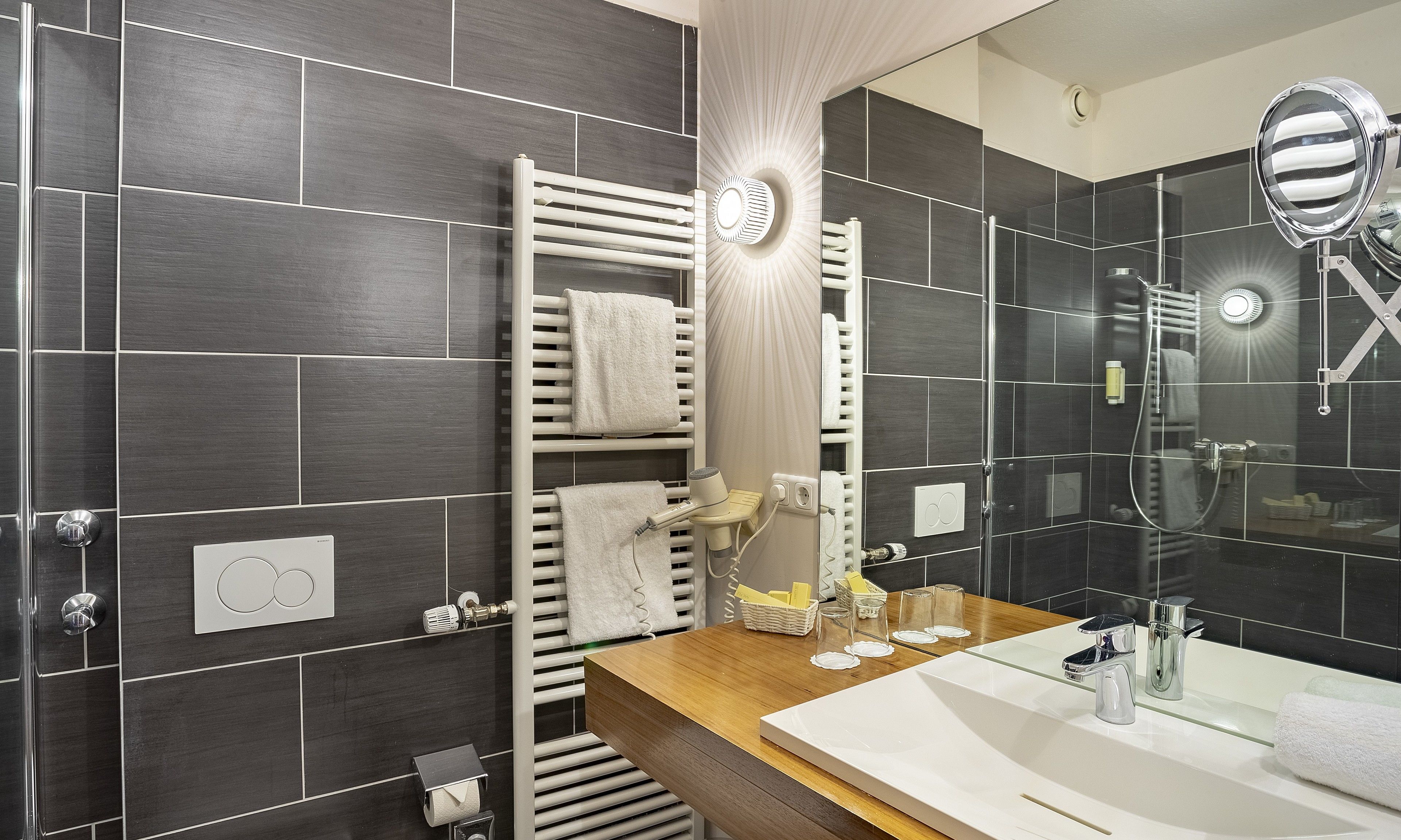A modern bathroom with dark tiled walls, a wooden vanity, a large mirror, and a glass shower enclosure.