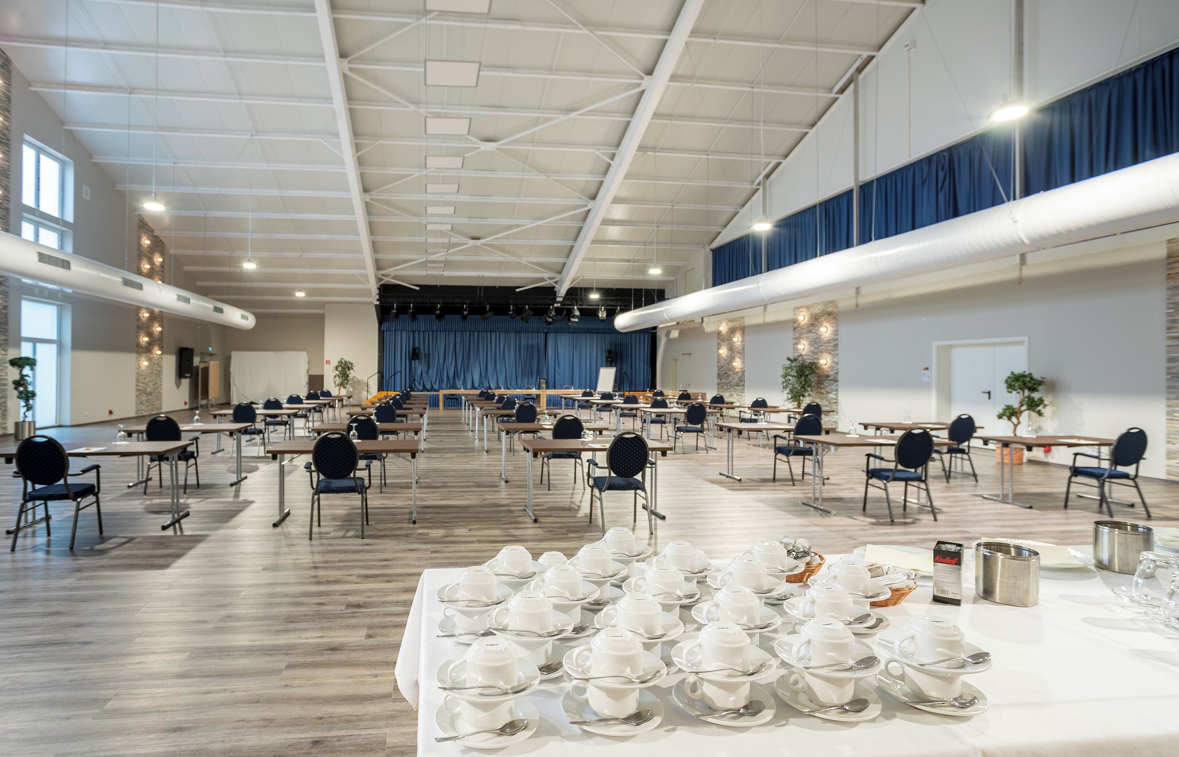 Un salón amplio con mesas dispuestas para una comida, que incluye un escenario y zona de estar.