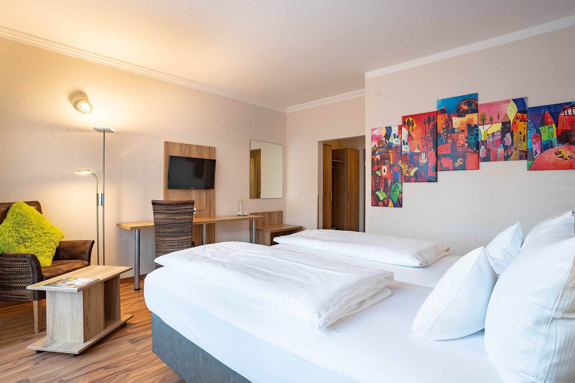 Ett hotellrum med en stor säng, ett skrivbord, en stol, en tv och en vägg prydd med färgstarka konstverk.