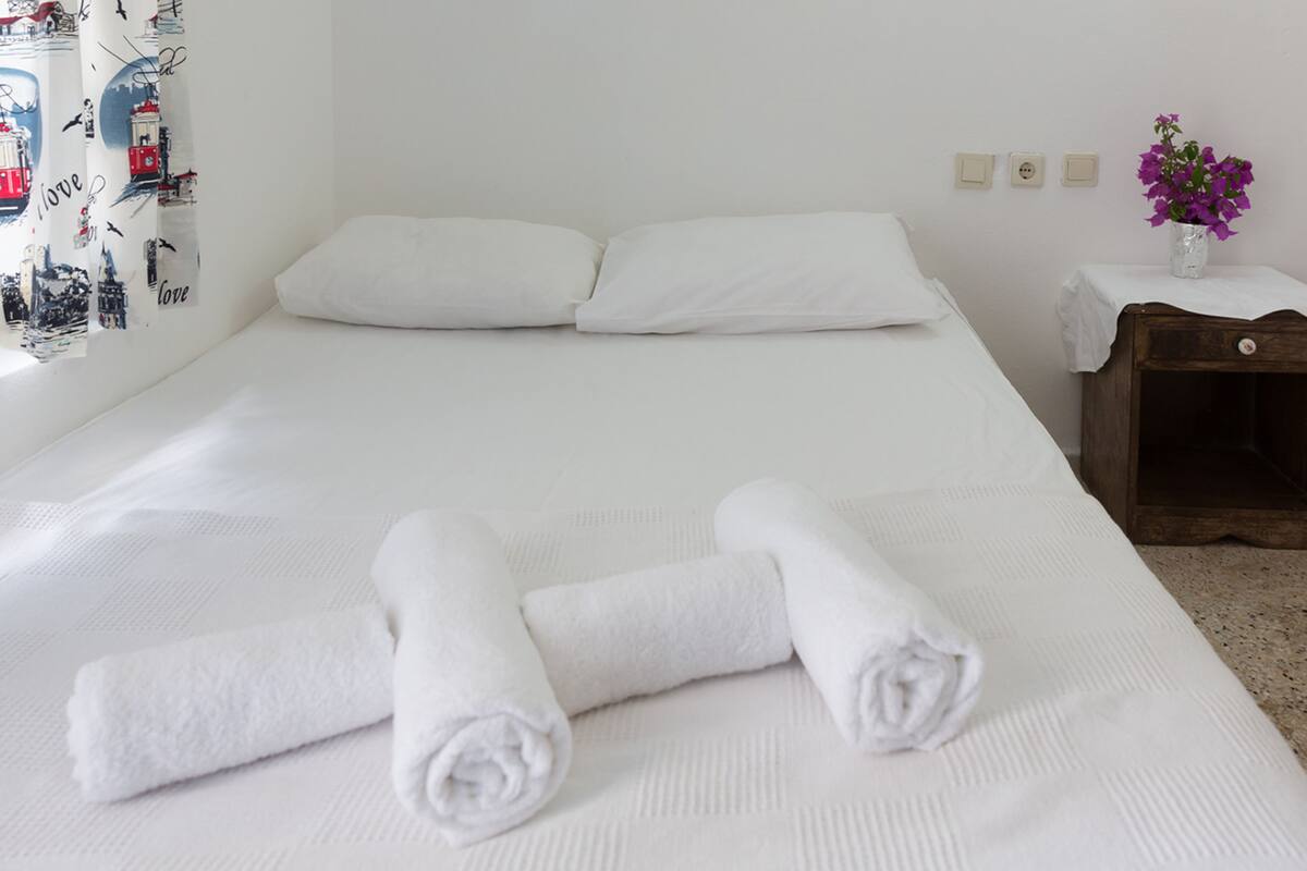 Una cama bien hecha con sábanas blancas y dos toallas enrolladas. A la derecha, una mesita de madera con un jarrón de flores moradas.