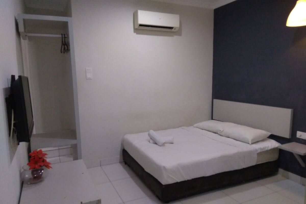 A small, clean room with a bed, a white desk, a TV, and an air conditioning unit.