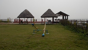 Uma área ao ar livre com gramado, equipamentos de exercício, duas treliças com telhado de palha e uma cerca.