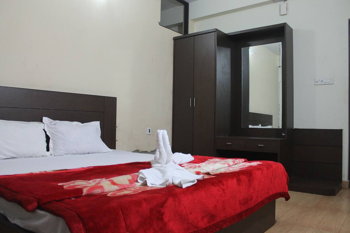 A hotel room with a bed, a red blanket, white pillows, a dark wooden wardrobe, and a mirror.