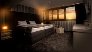 Een moderne slaapkamer met een groot bed, nachtkastjes, een bureau en uitzicht op een zonsondergang door het raam.