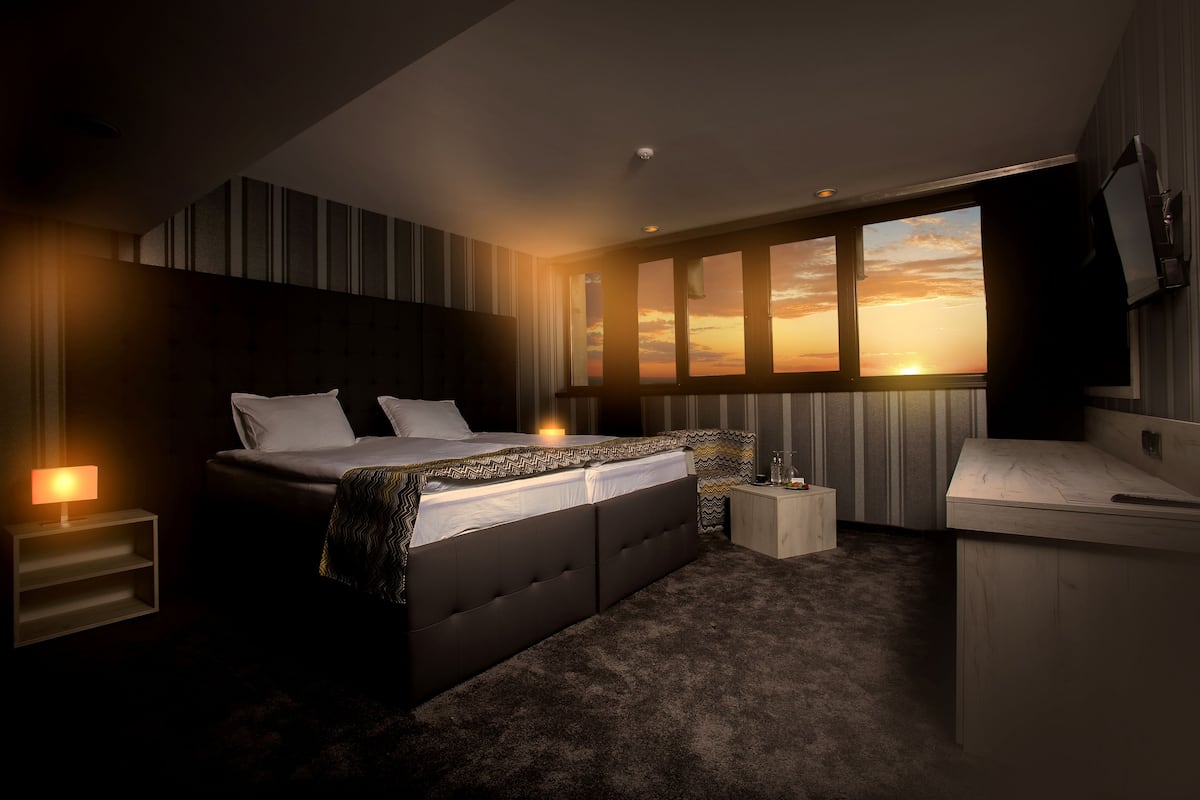 A modern bedroom with a large bed, bedside tables, a desk, and a view of a sunset through the window.