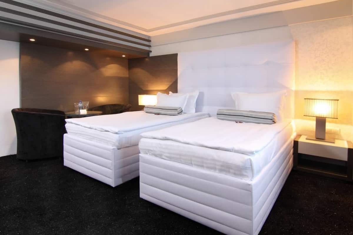 A hotel room with two beds, a dark carpet, a nightstand with a lamp, and a wall with a textured pattern.