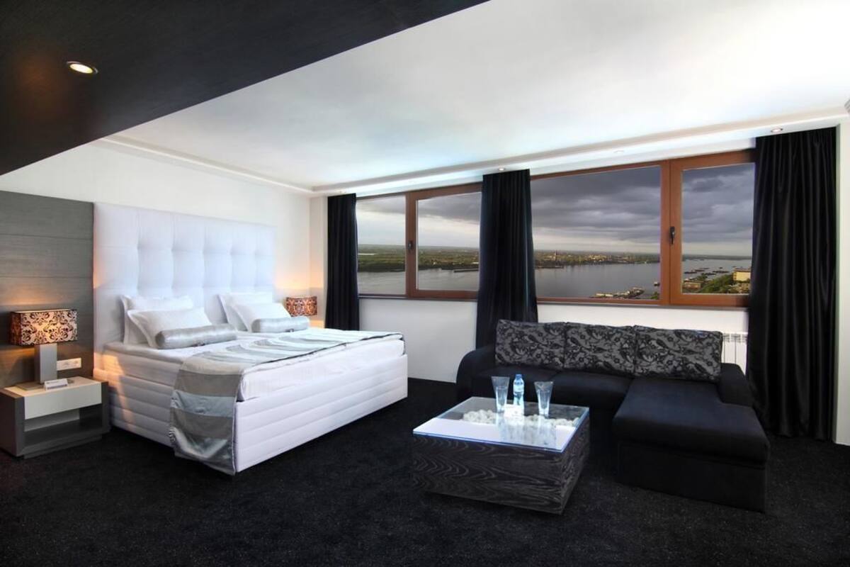 A modern hotel room with a large bed, a sofa, and a coffee table. There is a view of a body of water through the window.