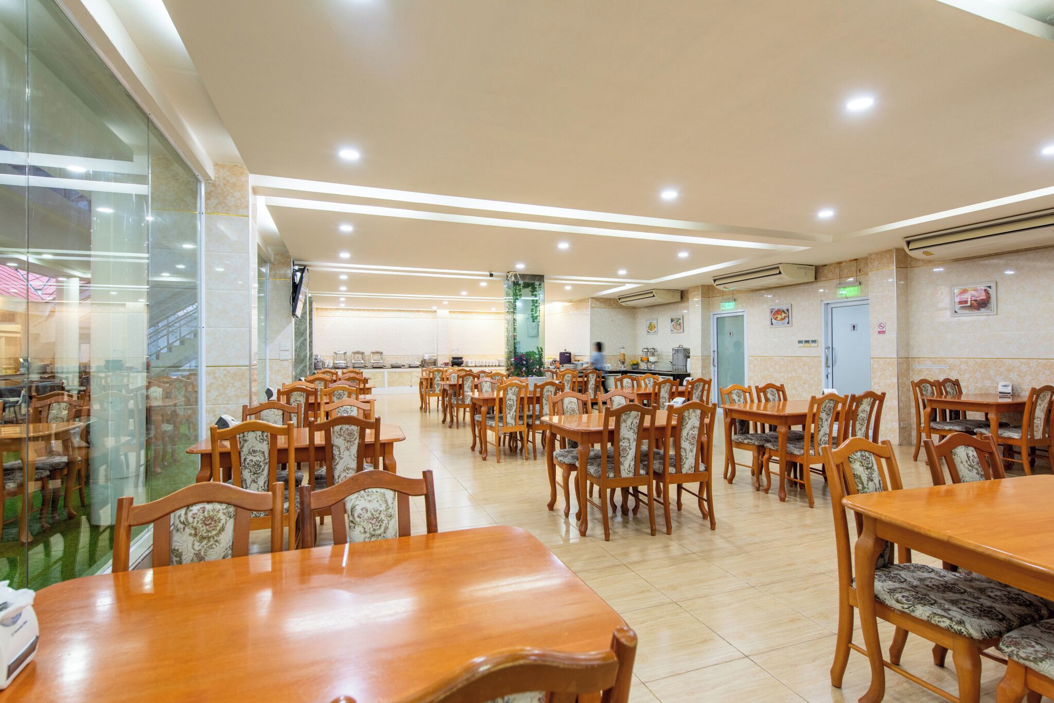 Un grand espace dédié à la salle à manger, avec de nombreuses tables et chaises en bois, un mur en verre d’un côté et un sol carrelé.