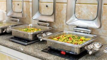 Un buffet composé de plusieurs plateaux de service en acier inoxydable contenant divers plats, d’une plaque de cuisson à induction intégrée et d’un pare-feu carrelé.