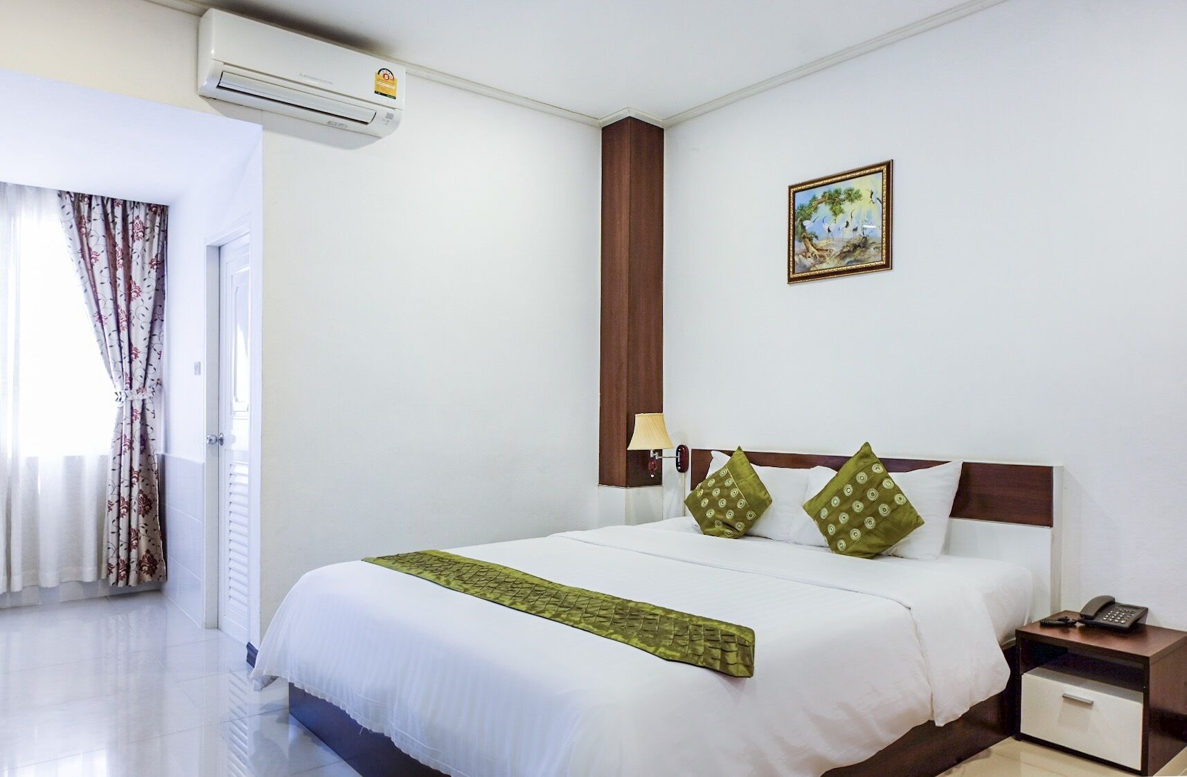 A hotel room with a bed, a nightstand, a lamp, a painting on the wall, and an air conditioning unit.