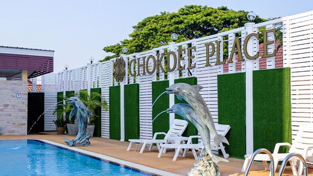 Zona de piscina con un cartel que dice "Chokdee Place", dos esculturas de delfines y sillas reclinables blancas.
