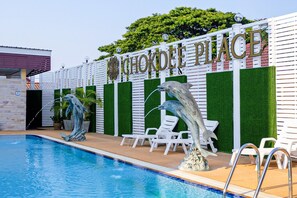 Zona de piscina con un cartel que dice "Chokdee Place", dos esculturas de delfines y sillas reclinables blancas.