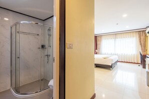 Una habitación de hotel moderna con ducha de vidrio, cama con ropa blanca y un gran ventanal con cortinas.