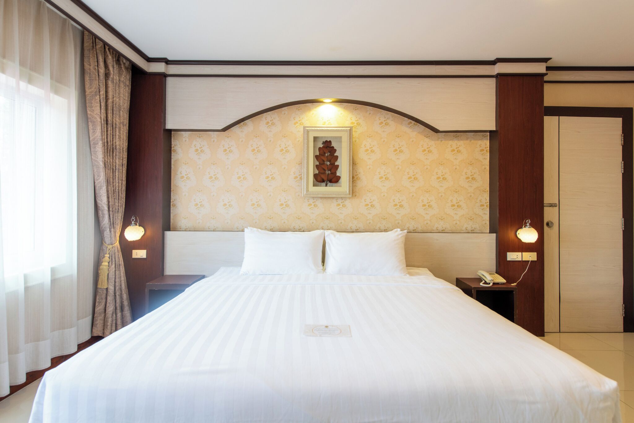 Ein Hotelzimmer mit einem großen Bett, einem Kopfteil mit dekorativer Zarge, zwei Nachttischlampen, einem Nachttischtelefon und einer Tür mit Lichtschalter.