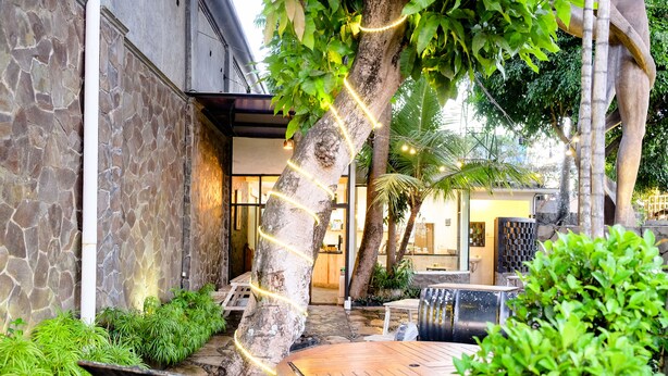 A stone-walled outdoor area with a wooden table and chairs, surrounded by greenery and string lights. - Pendhapa Art Space - PAS Limasan Homestay (Sewon)