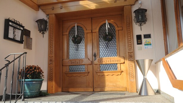 A wooden double door with "Hotel" and "Raiffeisen" signs, a potted plant, and a metal funnel on the steps. - Hotel Reischenau (Ustersbach)