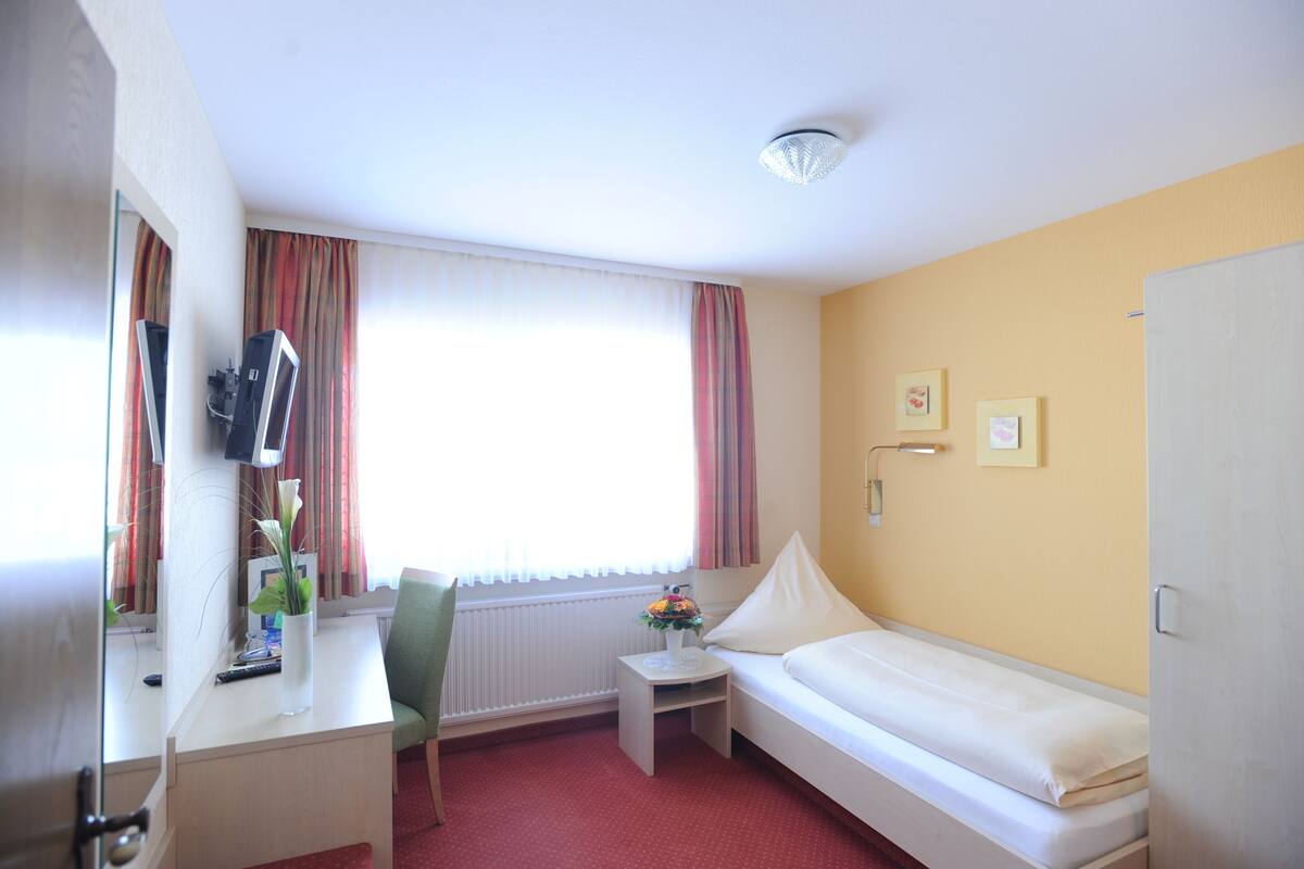 A hotel room with a bed, a desk, a chair, a small table with a vase of flowers, a television, and a window with curtains.