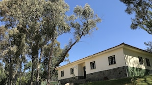 A two-story building with a stone foundation and a flat roof, surrounded by trees and a stone retaining wall.