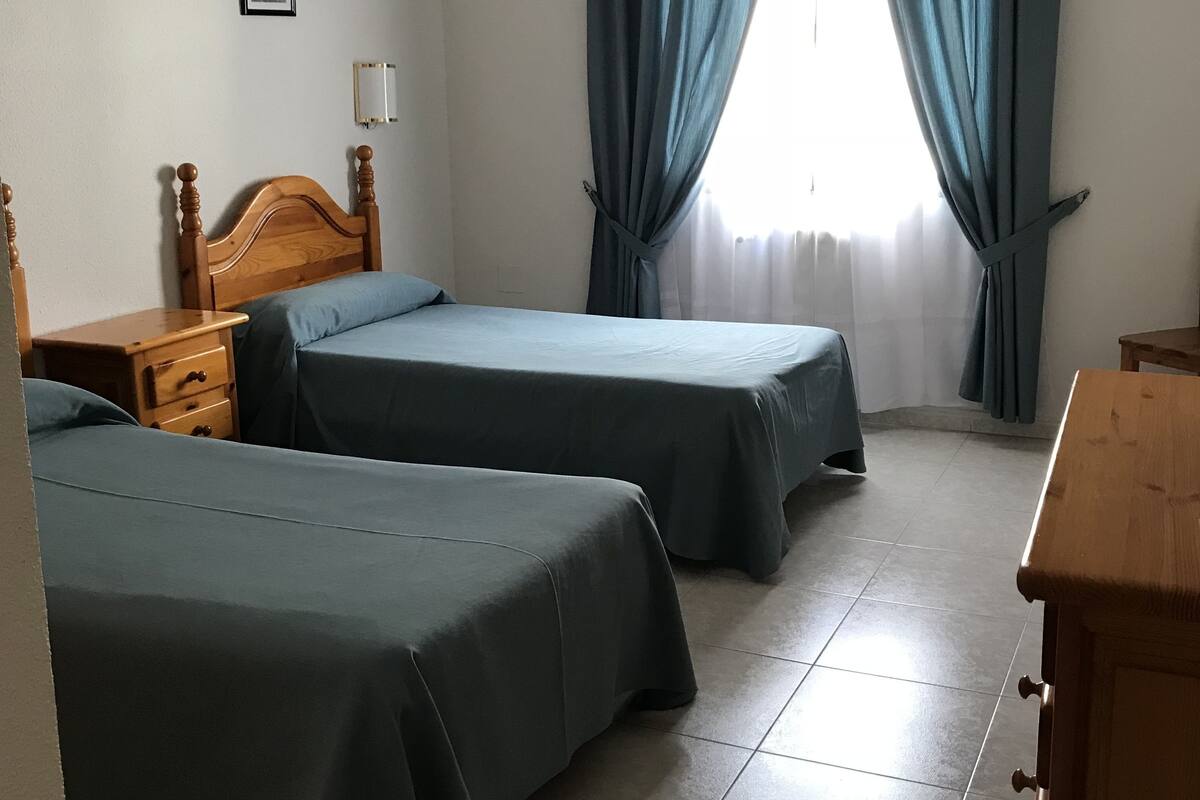 Una habitación con dos camas, un aparador de madera, una mesita de noche y una lámpara de pared.
