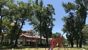 A playground with a slide and climbing structures, surrounded by trees and a building with a red roof.