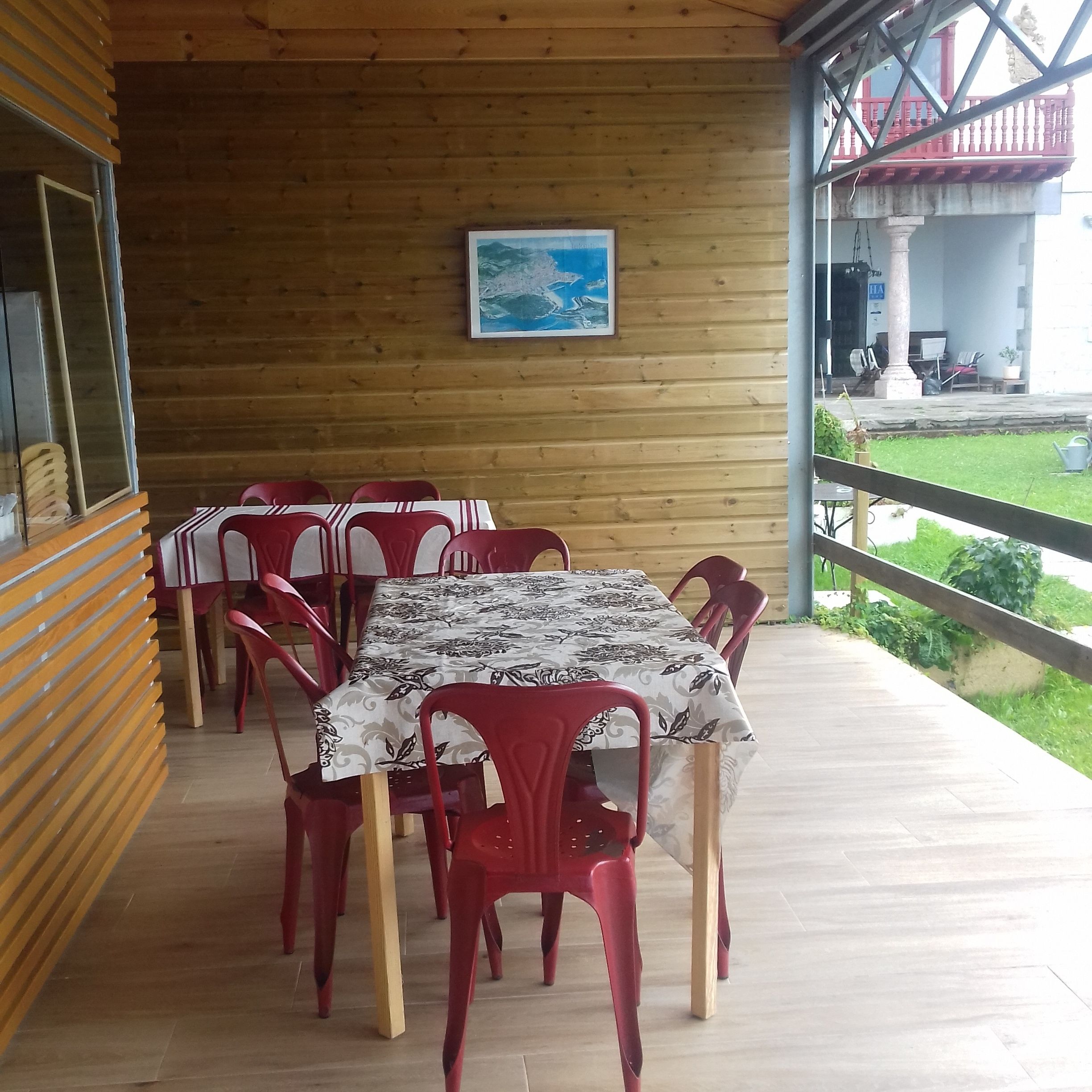 A covered outdoor dining area with wooden walls, a table with a patterned tablecloth, and red plastic chairs.