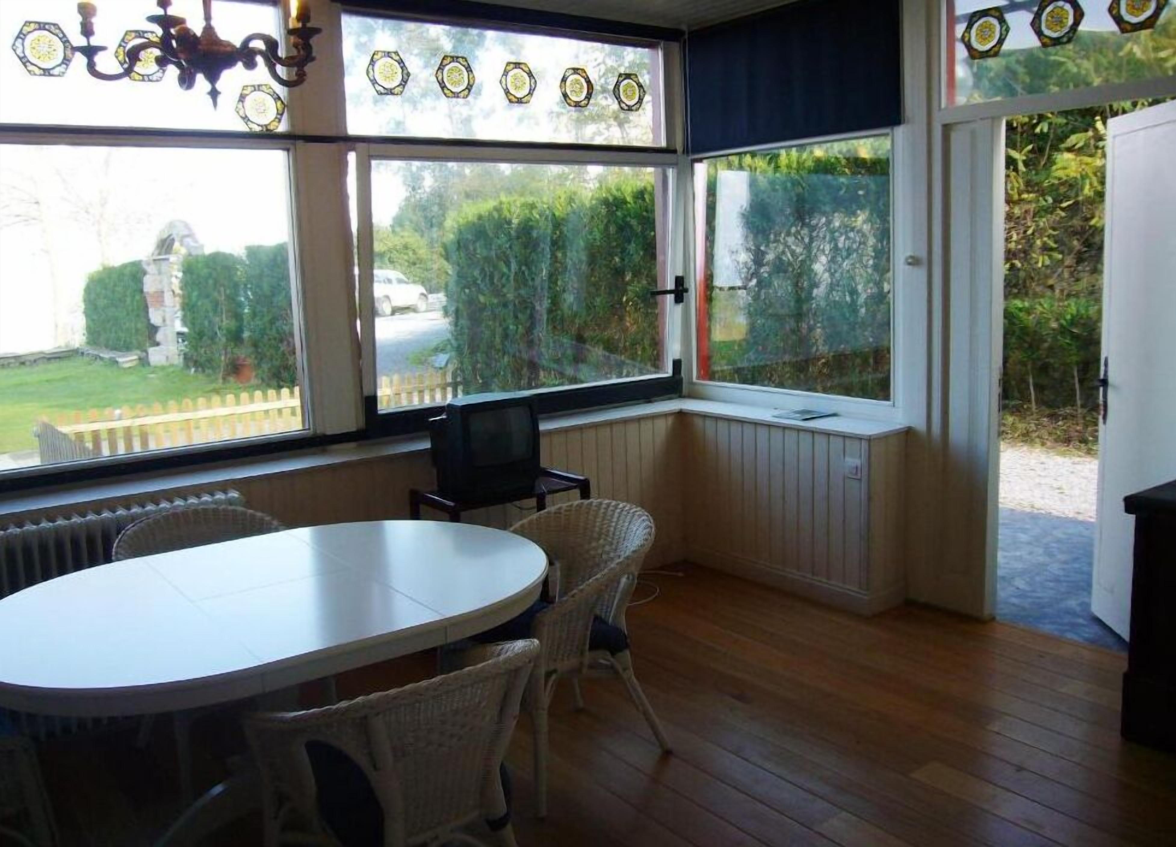 A room with a round table and wicker chairs, large windows with a view of greenery, and a radiator under the windows.