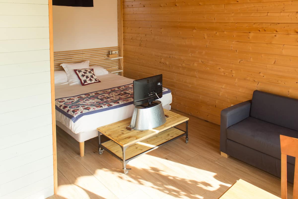 A bedroom with a bed, a wooden table with a kettle, a gray sofa, and a wooden wall.