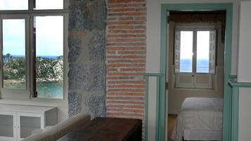 A room with a stone wall, a wooden bench, a bed, and a view of the sea through a window.