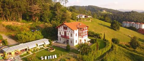 A traditional half-timbered house with a red-tiled roof, surrounded by lush greenery and hills.