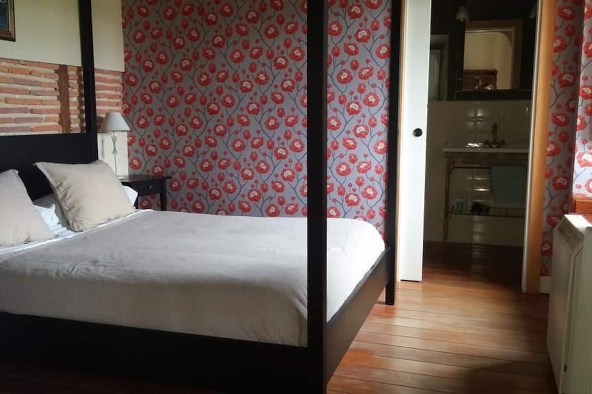 A bedroom with a four-poster bed, a brick accent wall, and a floral-patterned wallpaper.