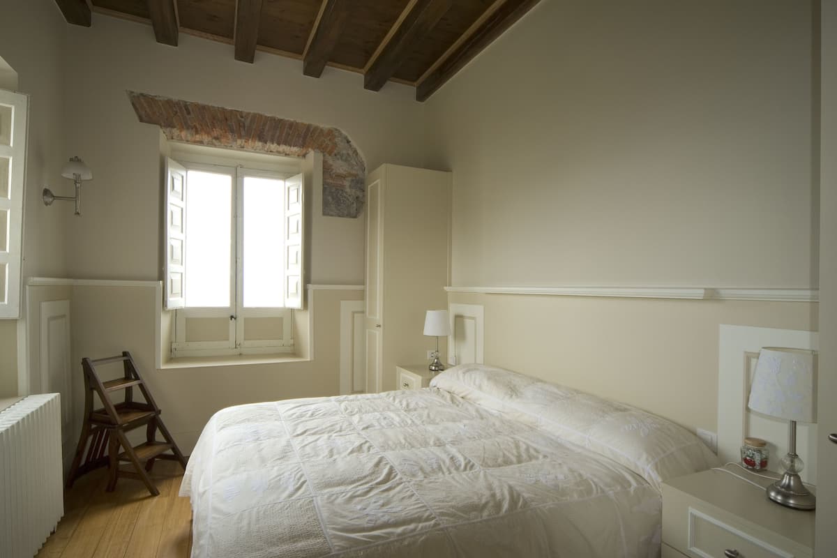 A bedroom with a bed, a window, a ladder, and a nightstand with a lamp.