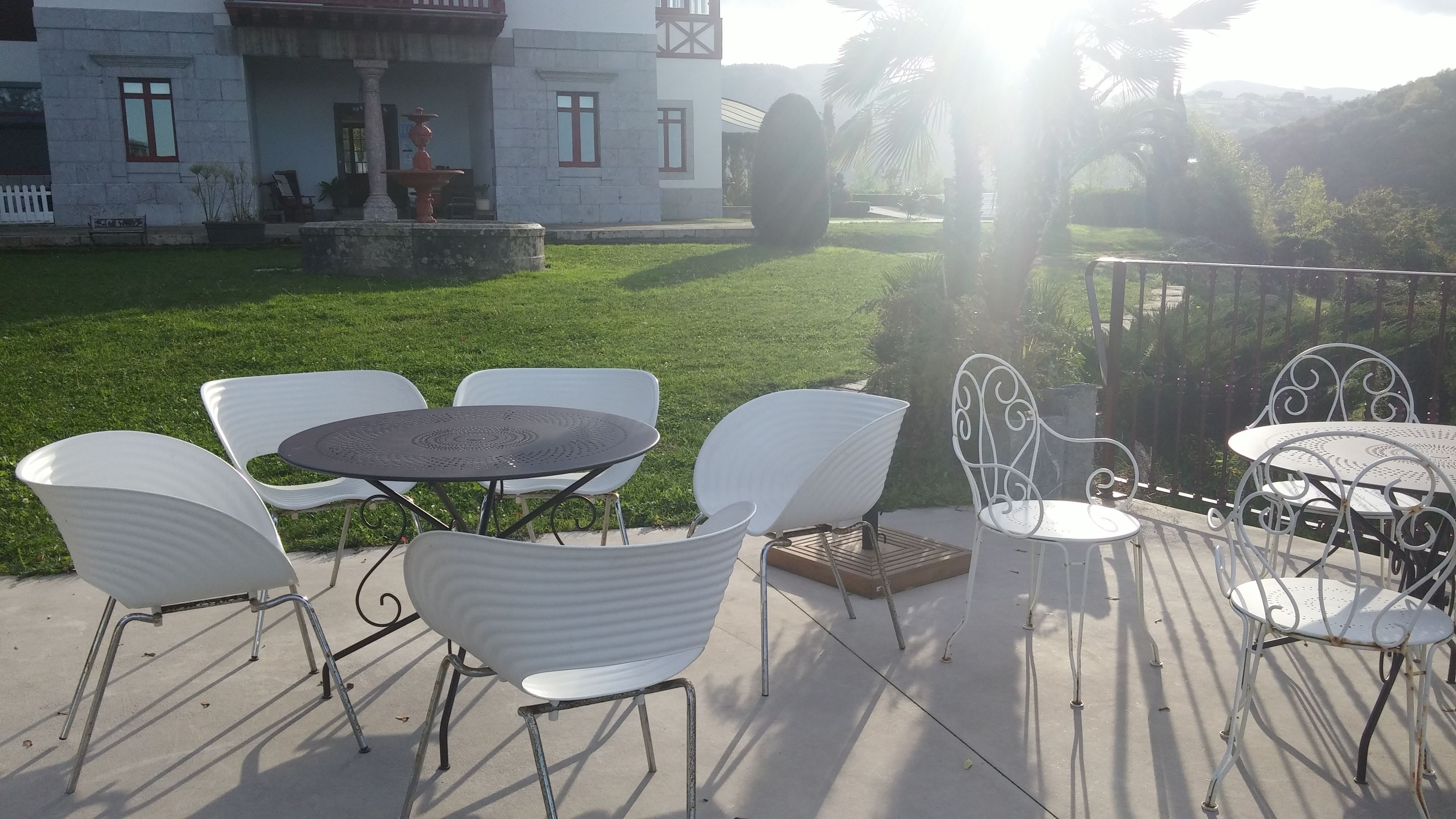 An outdoor patio with white metal chairs and tables, a stone fountain, and a building with a white facade and red window frames in the background.