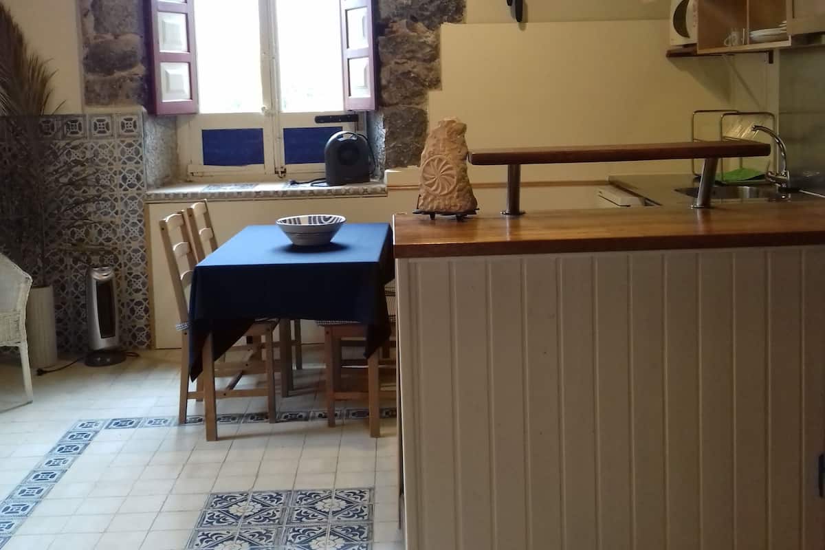 A kitchen with a stone wall, a window with shutters, a wooden table with a bowl, and a microwave on the counter.