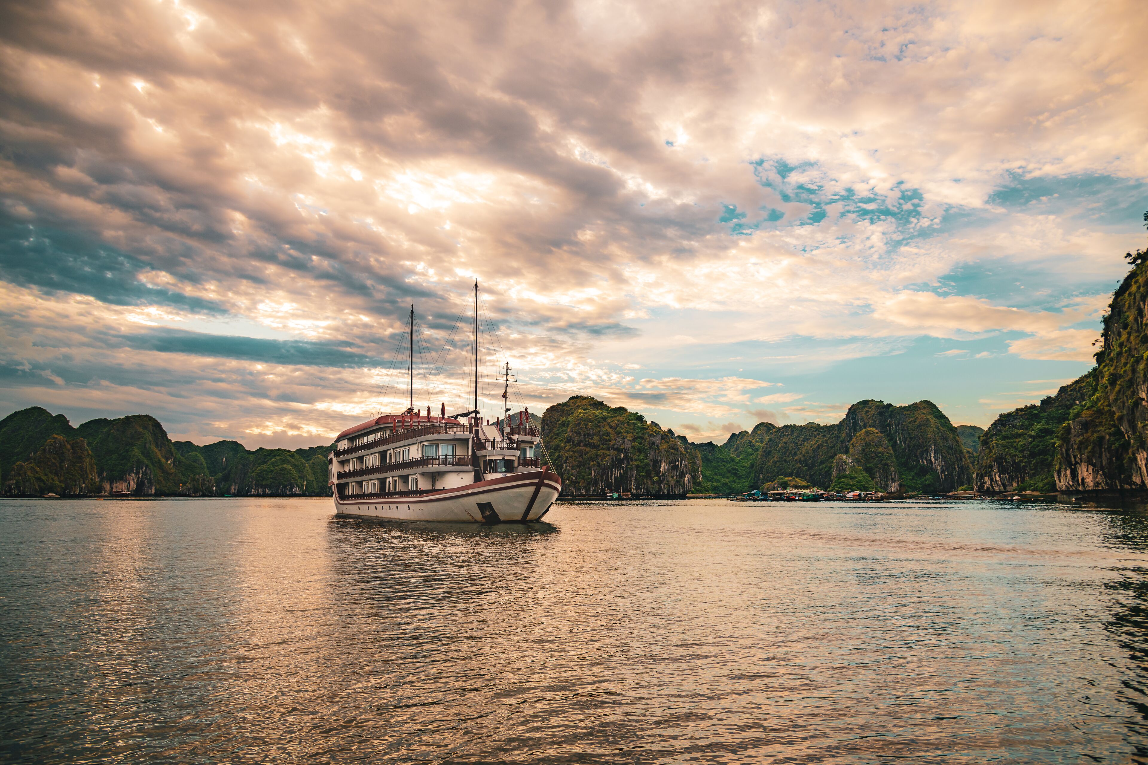 A boat with multiple decks and a red roof, floating on calm water with a backdrop of green hills and a cloudy sky.