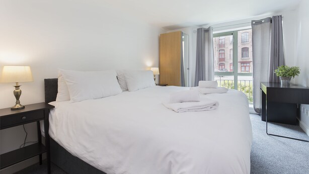 A hotel room with a neatly made bed, a bedside table with a lamp, a desk with a plant, and a window with curtains. - Raleigh Square Apartments (Nottingham)